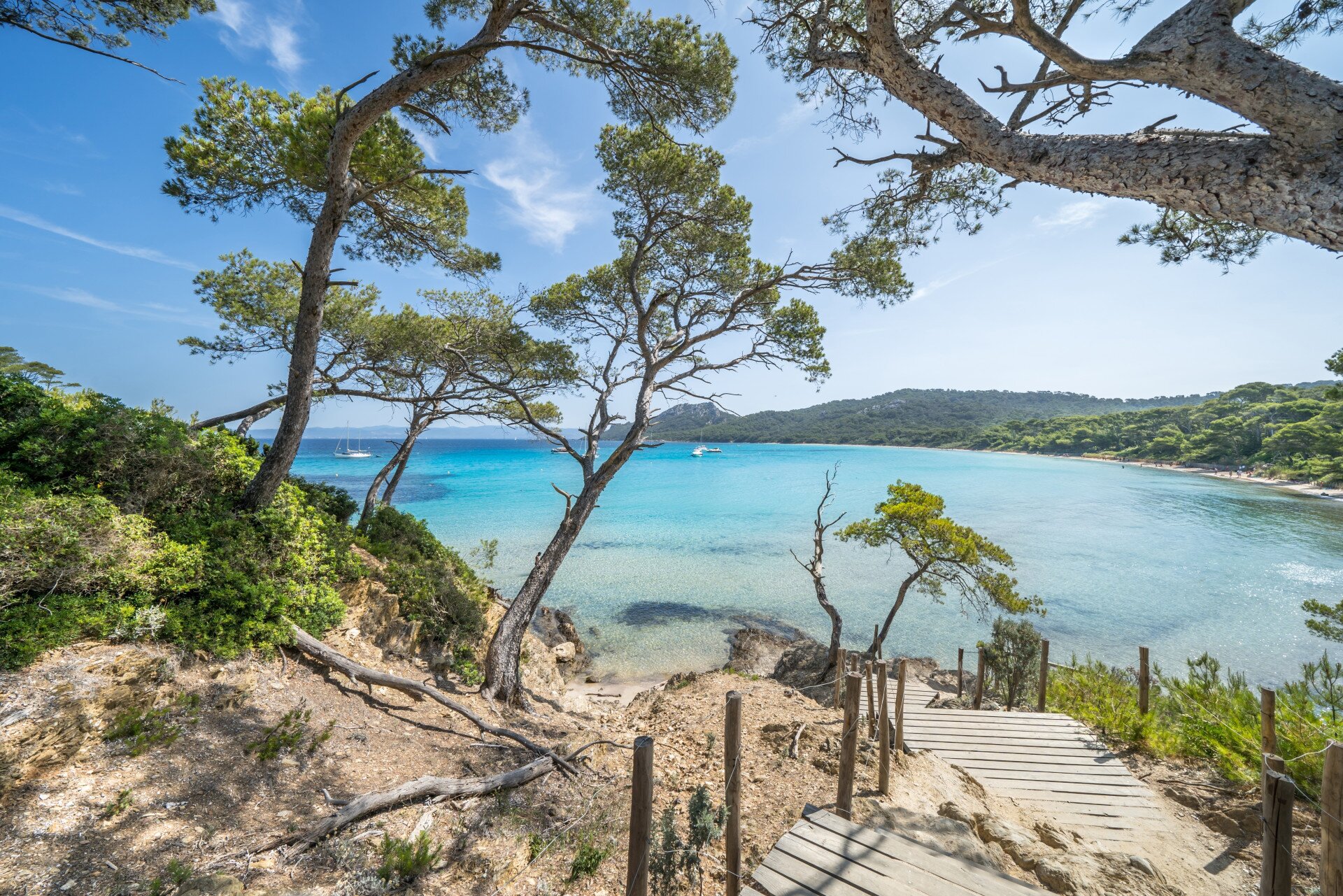 Wooden path leading down to a quiet turquoise bay framed by pine trees on the French Riveria in the winter, with clear skies and calm sea.