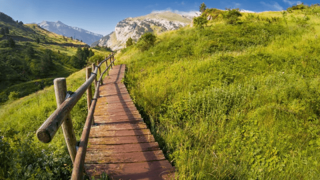 A wooden boardwalk in Saint-Jean-Pied-de-Port&nbsp;to Pamplona with a rustic railing winding through a lush green hillside, with mountains in the background under a bright blue sky.