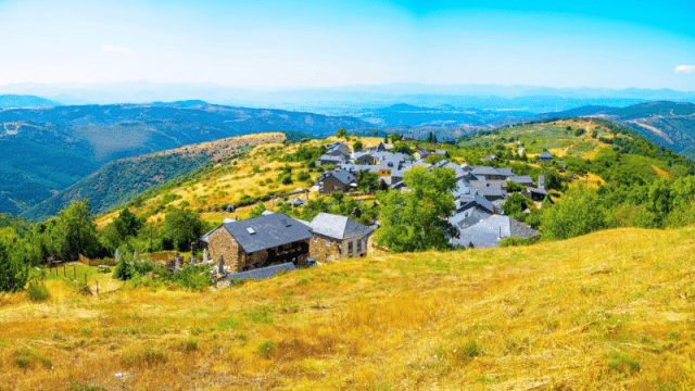 Sarria to Santiago de Compostela, a small mountain village with stone houses and slate roofs sits on a sunny hillside surrounded by rolling green and golden terrain, with expansive mountain ranges in the distance under a bright blue sky.